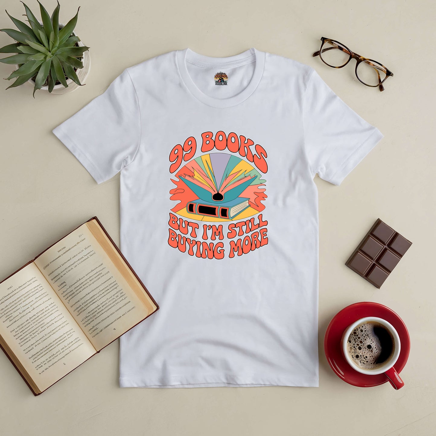 White t-shirt with "99 Books But I'm Still Buying More" design, surrounded by an open book, coffee, chocolate, plant, and glasses.