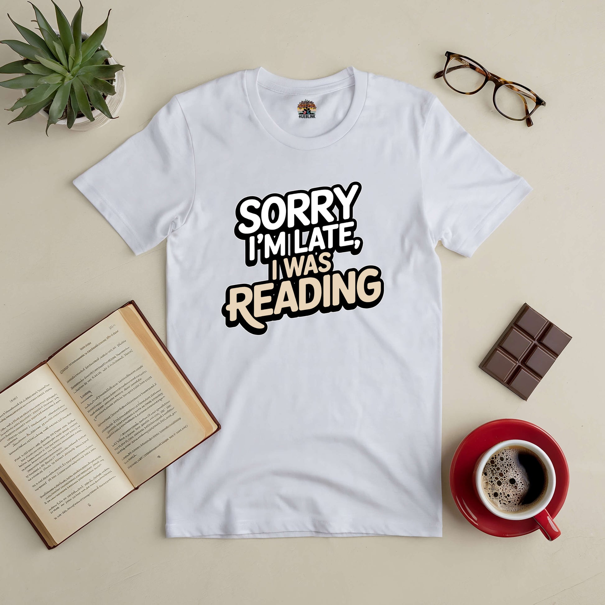 White tee with "Sorry I'm Late, I Was Reading" text, surrounded by glasses, open book, coffee, chocolate, and a plant, ideal for book lovers.