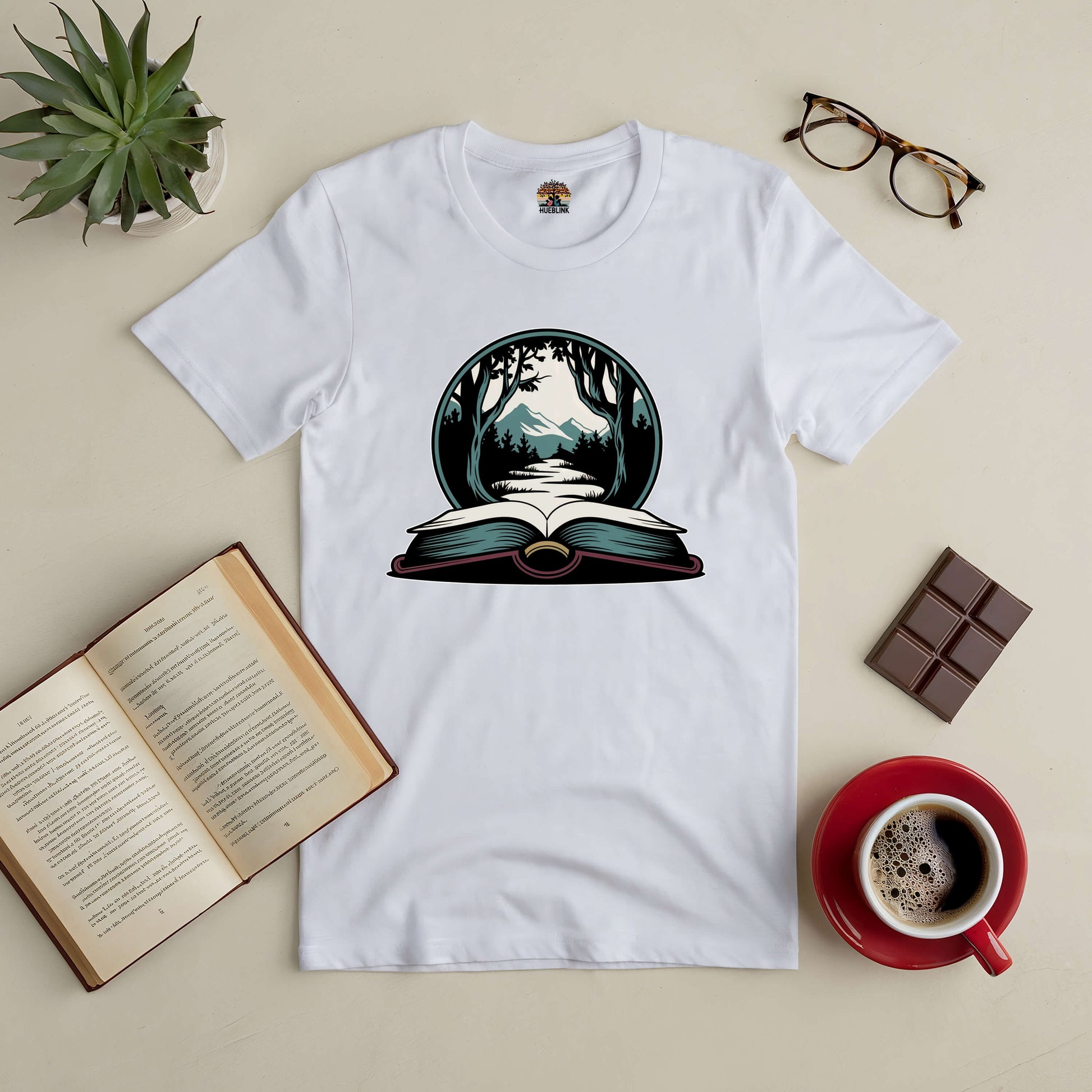 "Whispers Of The Wild Tee with nature-inspired book design, surrounded by books, coffee, plant, and glasses on a cozy table setting."