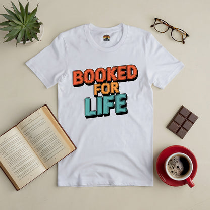 White tee with "Booked for Life" text, surrounded by a book, coffee, chocolate, glasses, and a plant on a beige surface.