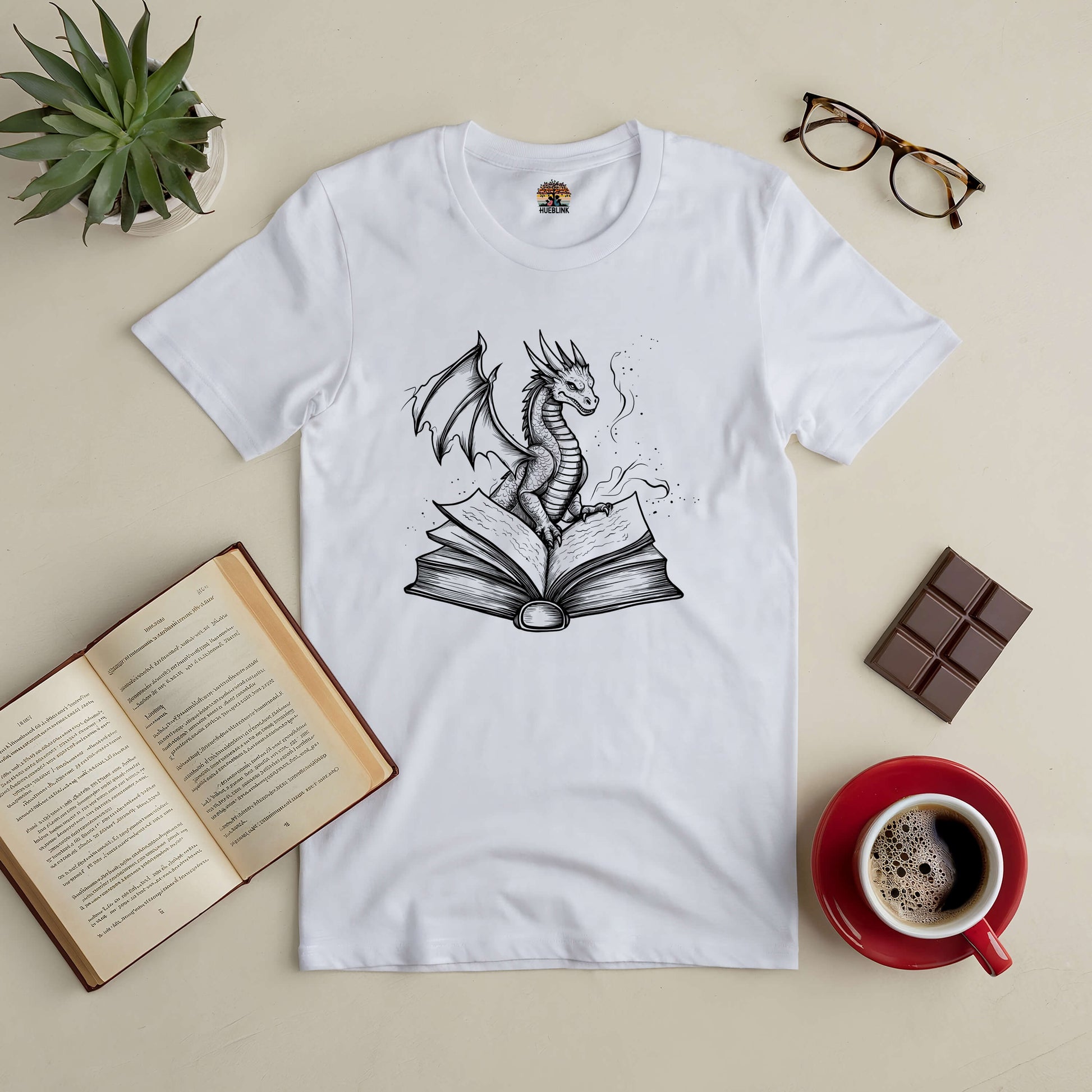 White t-shirt featuring a dragon emerging from a book, surrounded by an open novel, coffee cup, chocolate, glasses, and a plant.