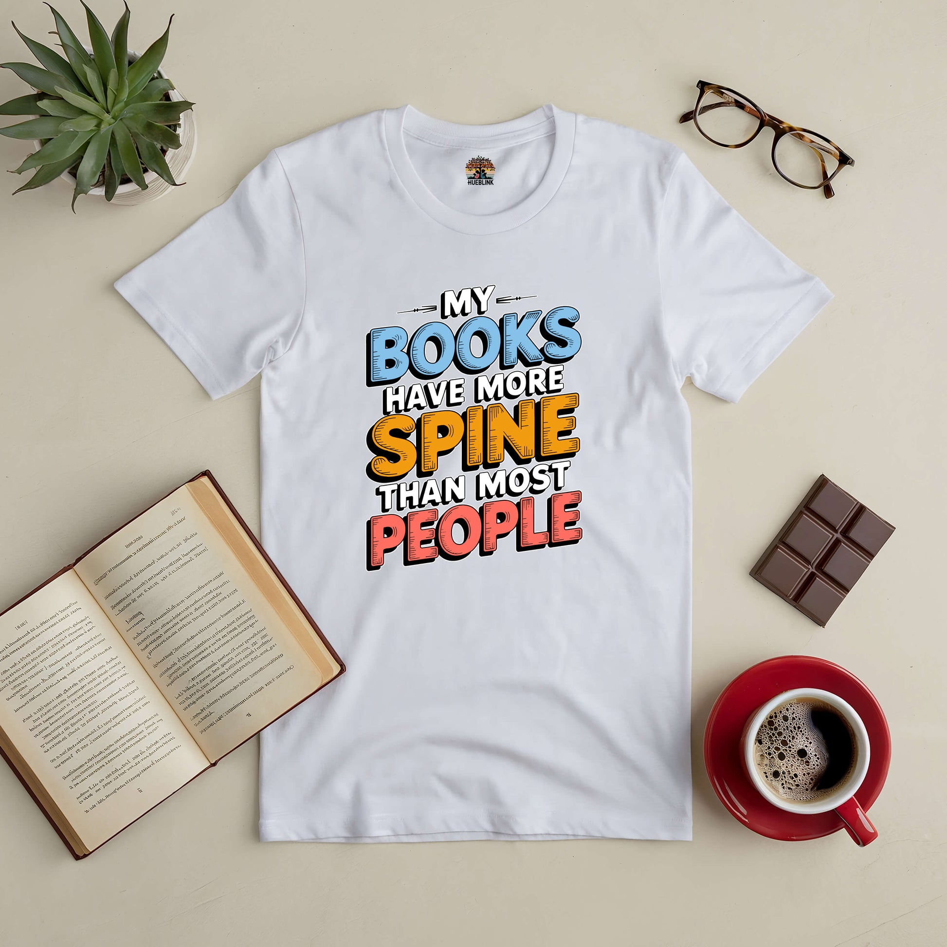 White t-shirt with "My Books Have More Spine Than Most People" text, surrounded by a book, glasses, chocolate, and coffee.