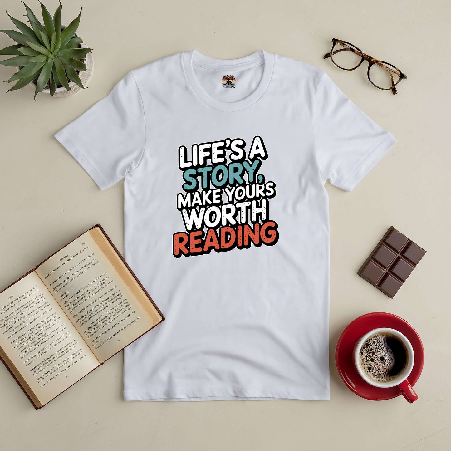 "Life's a Story Tee with motivational quote, surrounded by a book, glasses, chocolate, and coffee on a table"