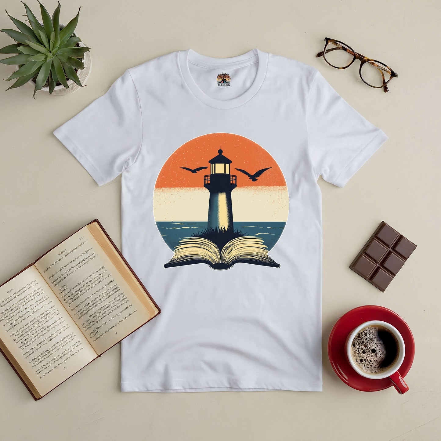 White tee with lighthouse and open book design, surrounded by coffee, open book, glasses, chocolate, and a plant on the table.