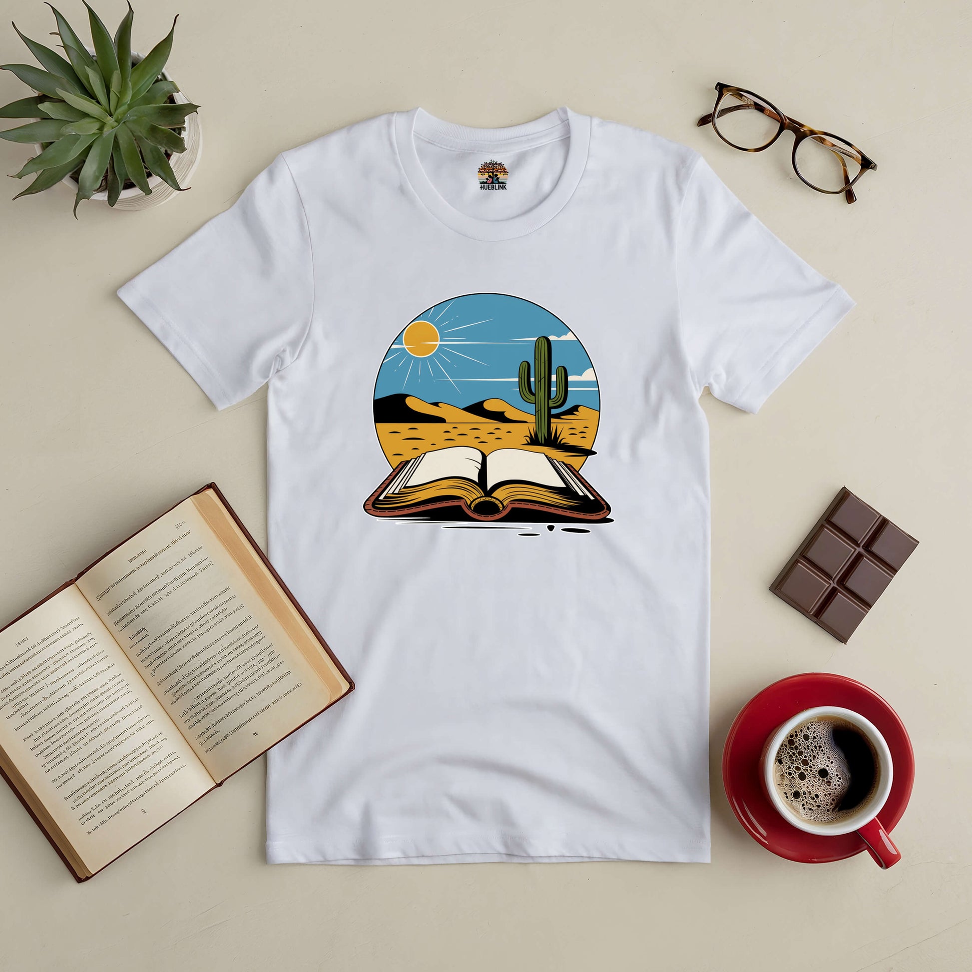 White Silent Desert Tee featuring desert and open book design, surrounded by coffee, chocolate, glasses, plant, and open book.