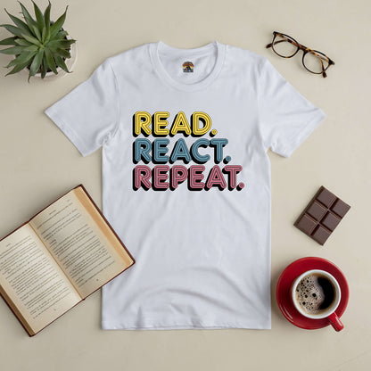 White tee with "Read. React. Repeat." text, surrounded by a book, glasses, chocolate, and a coffee cup on a table.