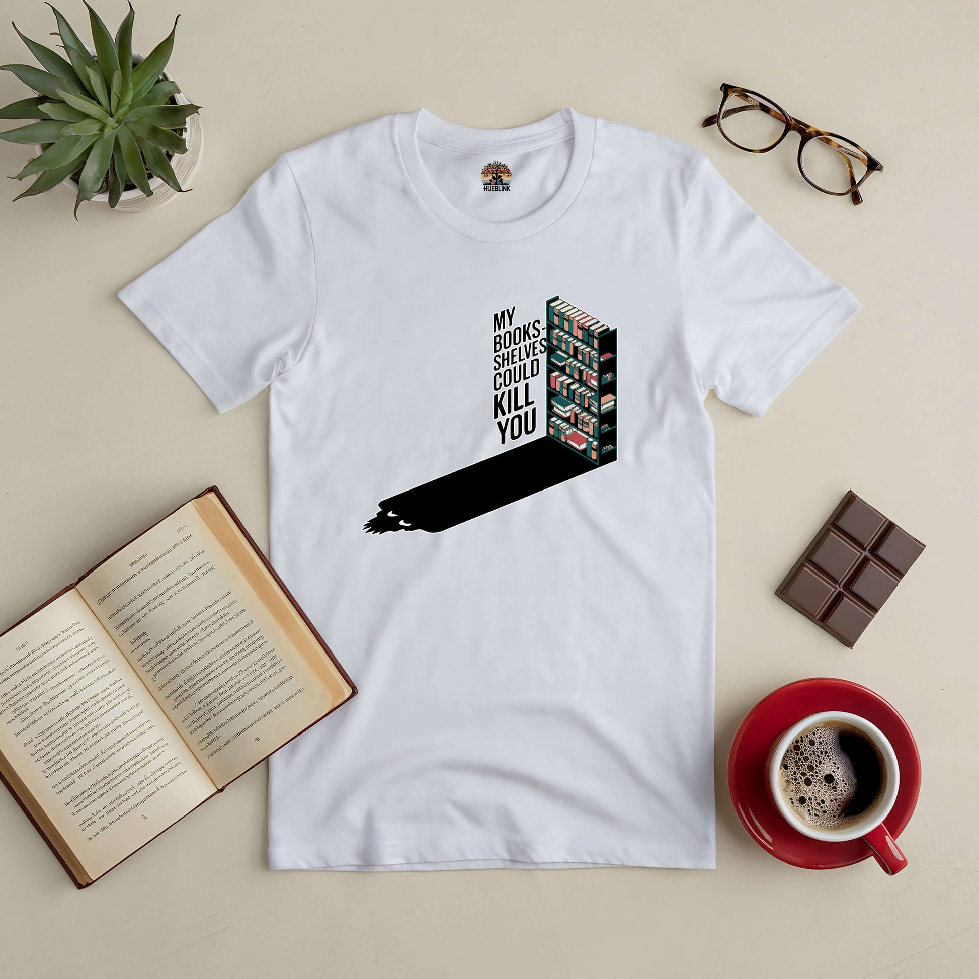 White tee with "My Bookshelves Could Kill You" graphic, surrounded by book, coffee cup, glasses, plant, and chocolate bar on a table.