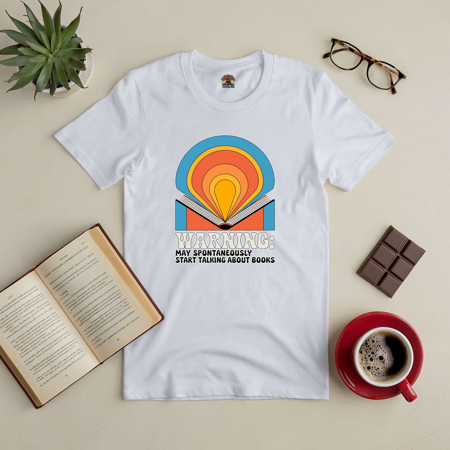 White tee with colorful book design and "Warning: May Spontaneously Start Talking About Books" text, surrounded by coffee and open book.