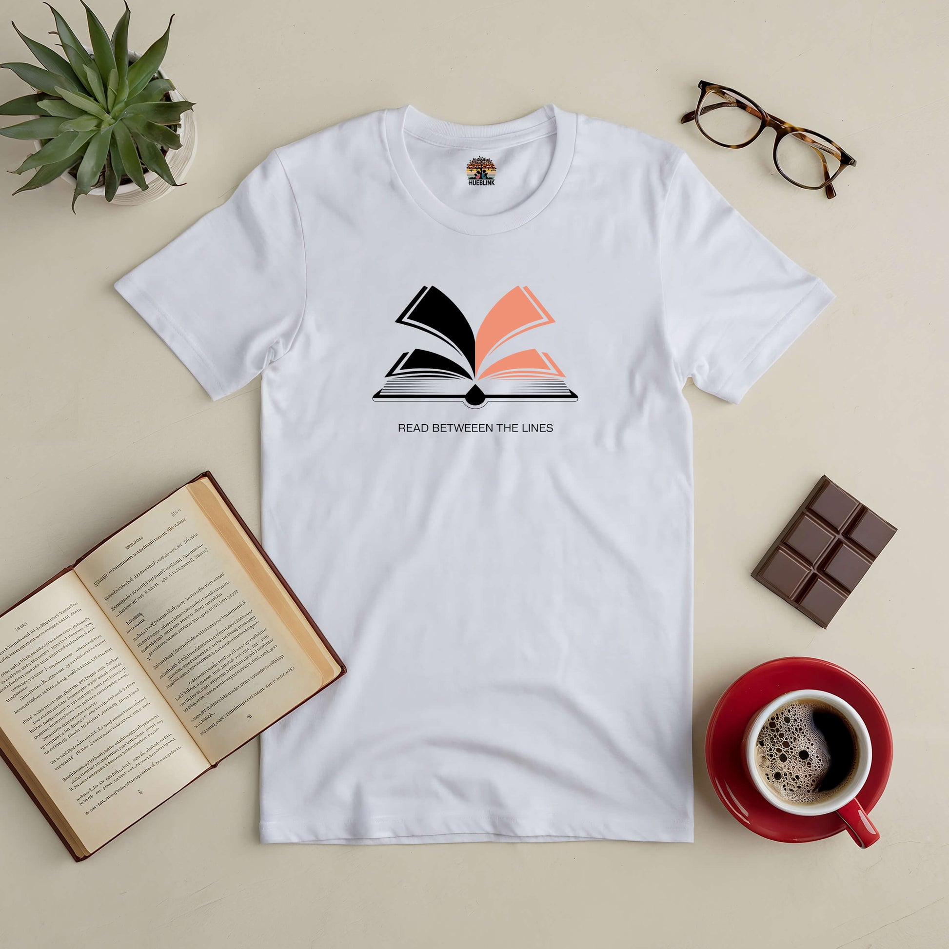 White "Read Between The Lines" tee with open book graphic, surrounded by a book, glasses, chocolate, and a coffee cup.