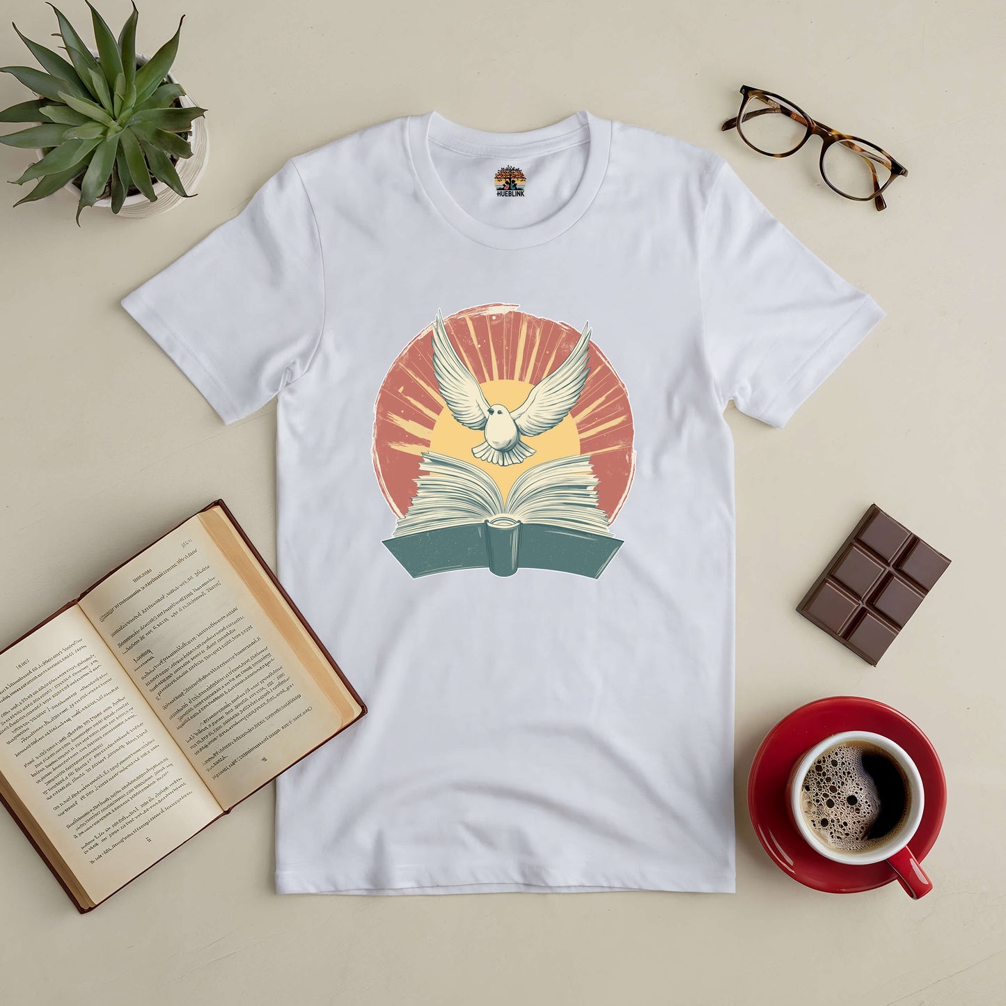 Boundless Horizons Tee with book and bird graphic, surrounded by coffee, chocolate, glasses, and open book on a table.