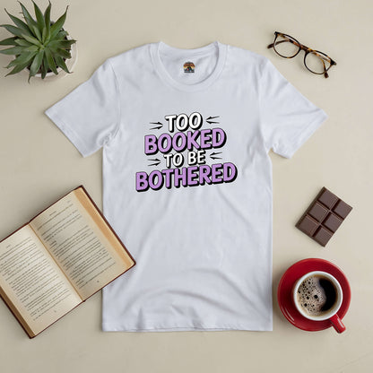 White "Too Booked to Be Bothered" tee on table with open book, coffee, chocolate, glasses, and plant.