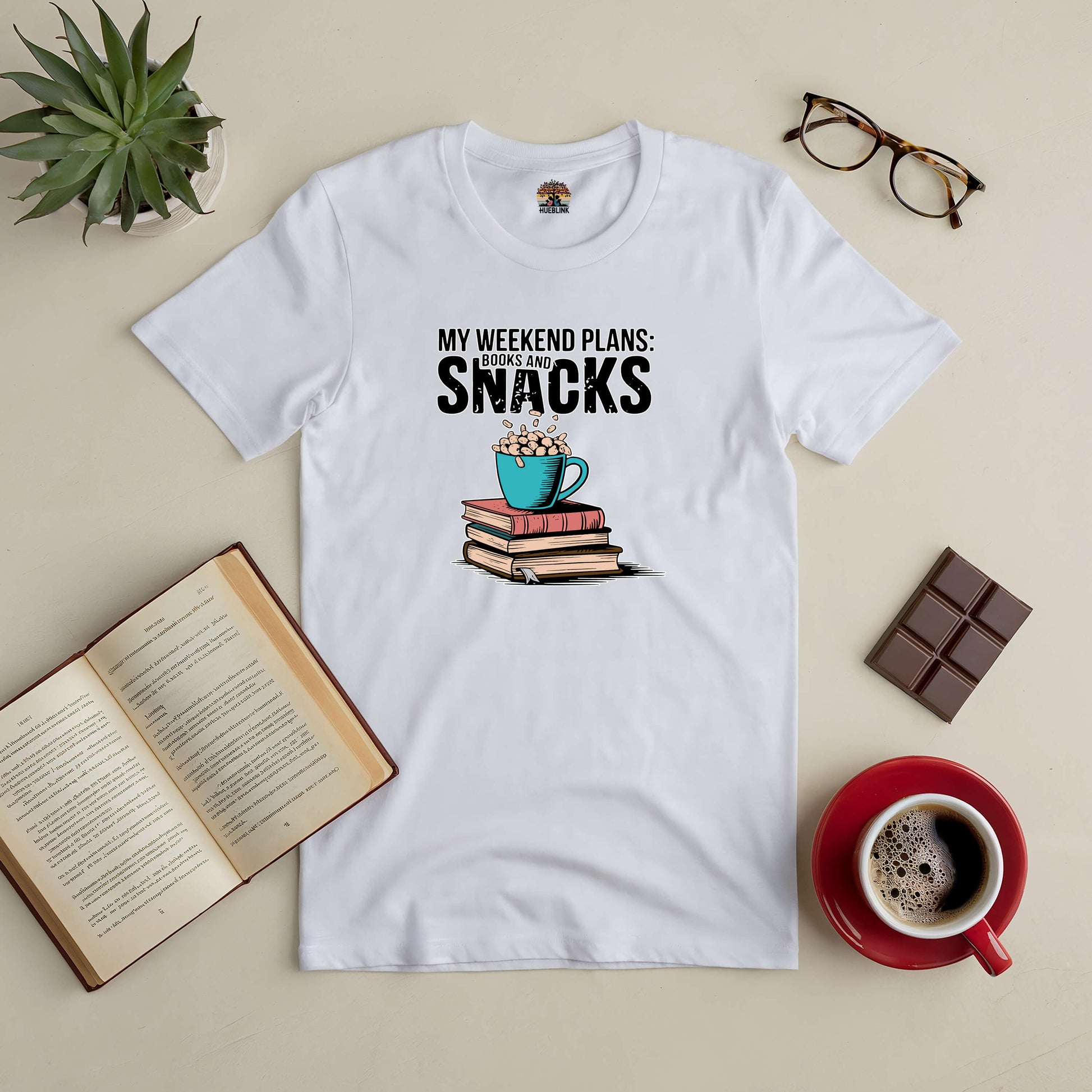 White tee with "My Weekend Plans: Books and Snacks" text, surrounded by a stack of books, a coffee cup, glasses, and chocolate.