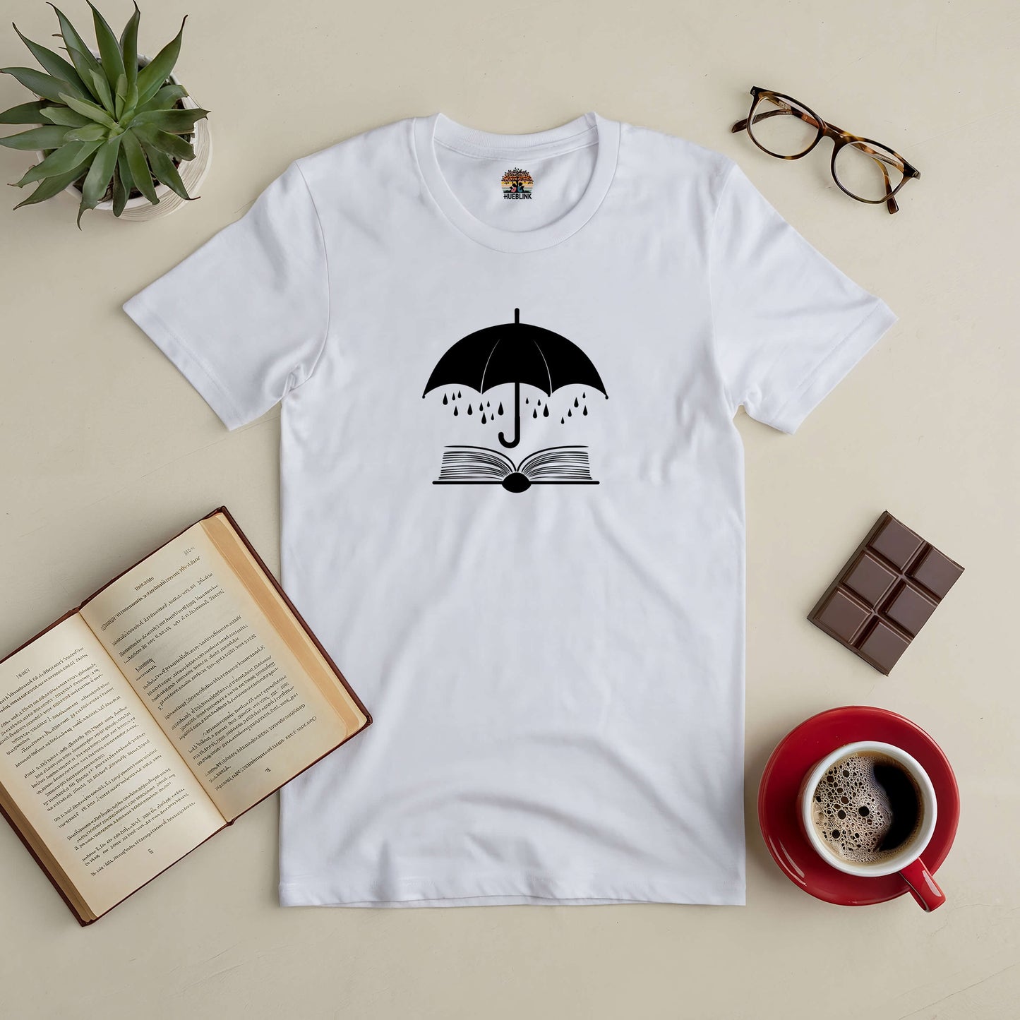 Sheltering Stories Tee with umbrella and book design, set on a table with open book, coffee, glasses, chocolate, and plant.