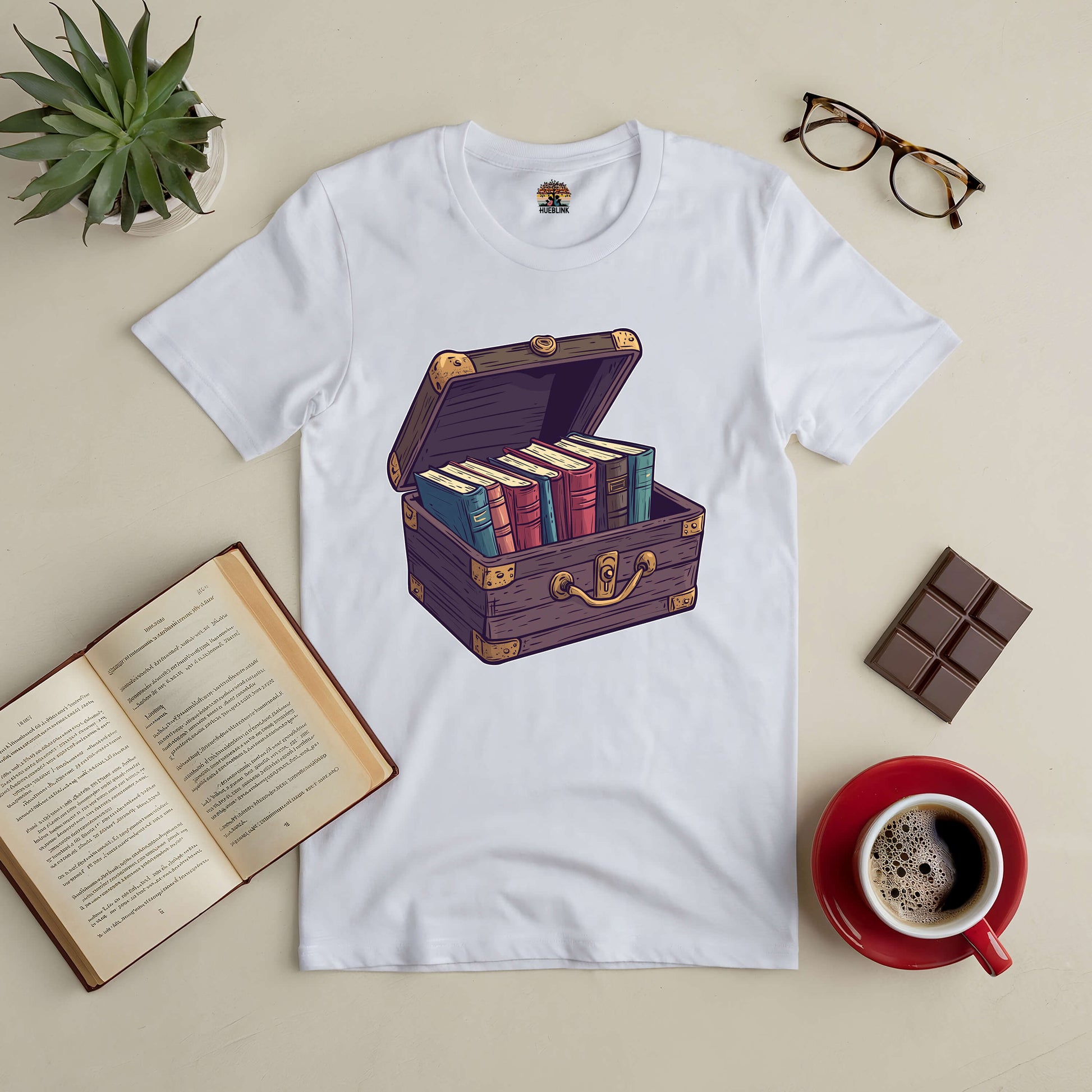 "Priceless Treasure Tee with book-filled chest design, surrounded by coffee, chocolate, and an open book on a table."