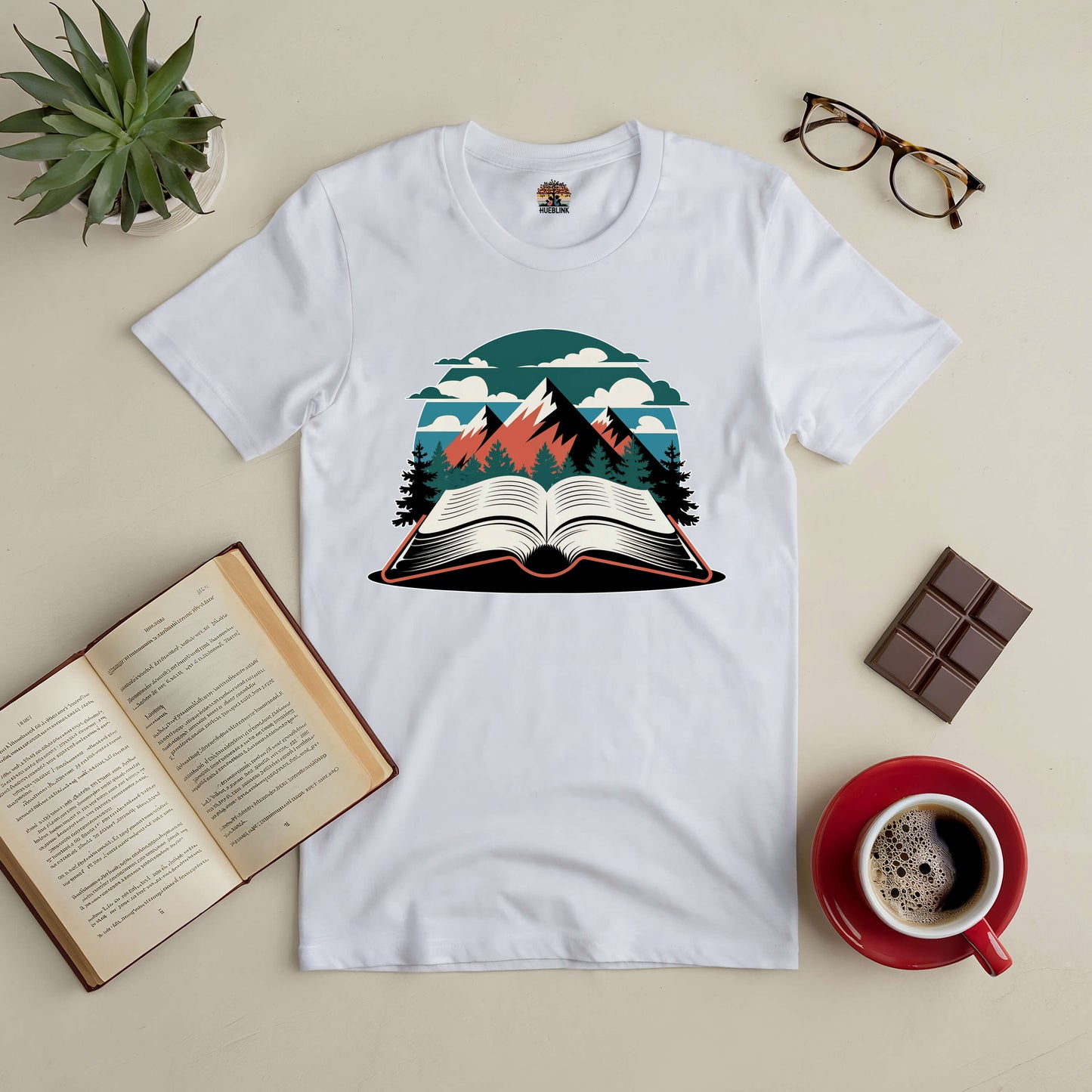 White tee featuring a mountain and open book design, surrounded by a book, glasses, chocolate, and coffee on a beige surface.