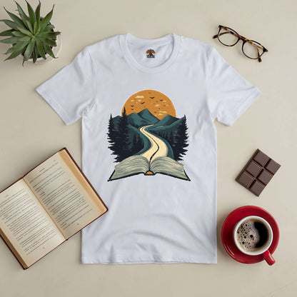 White tee with open book graphic leading to mountains and sunset, surrounded by coffee, glasses, chocolate, and succulents.