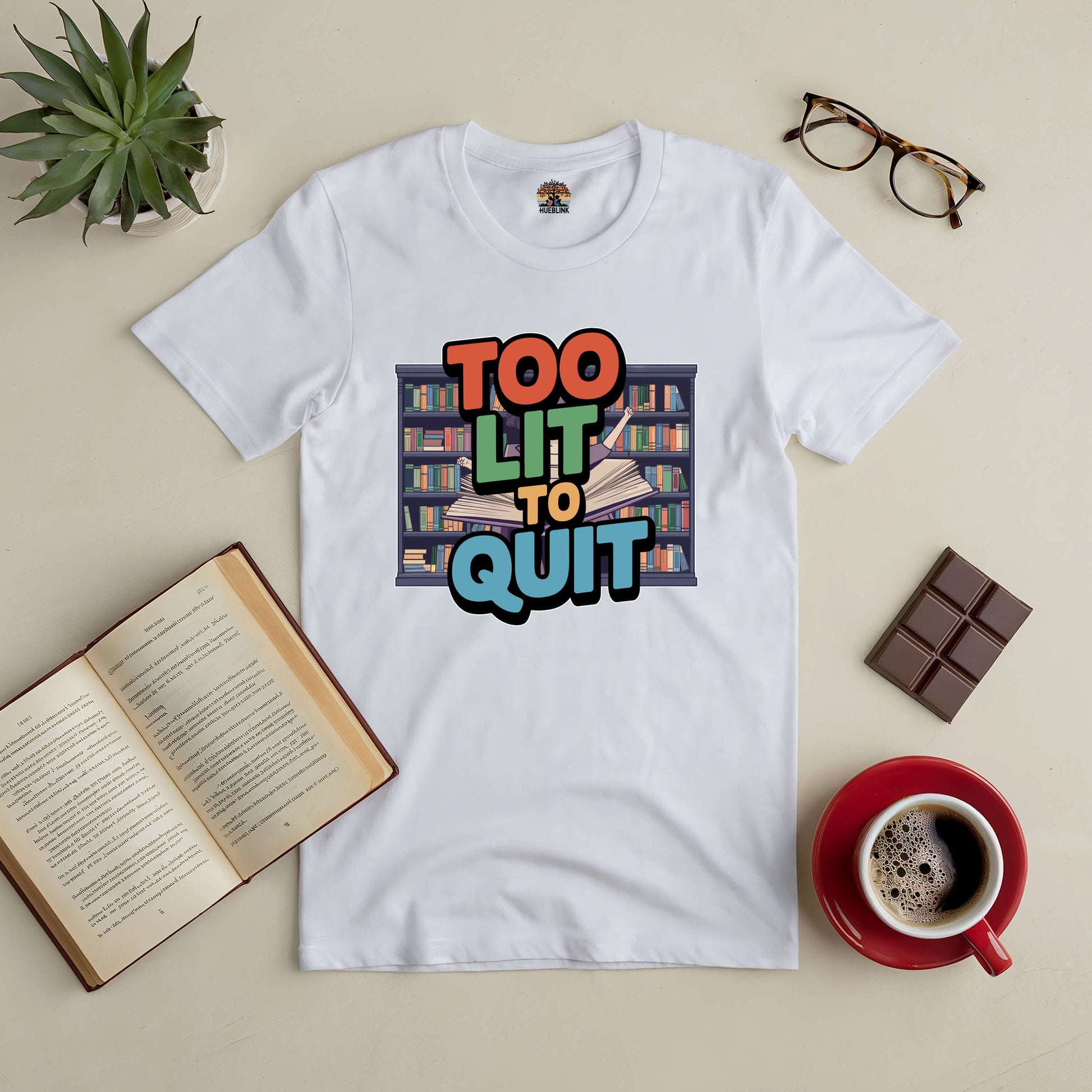 "Too Lit To Quit t-shirt featuring colorful bookshelf design, perfect for passionate book lovers, surrounded by coffee and books."