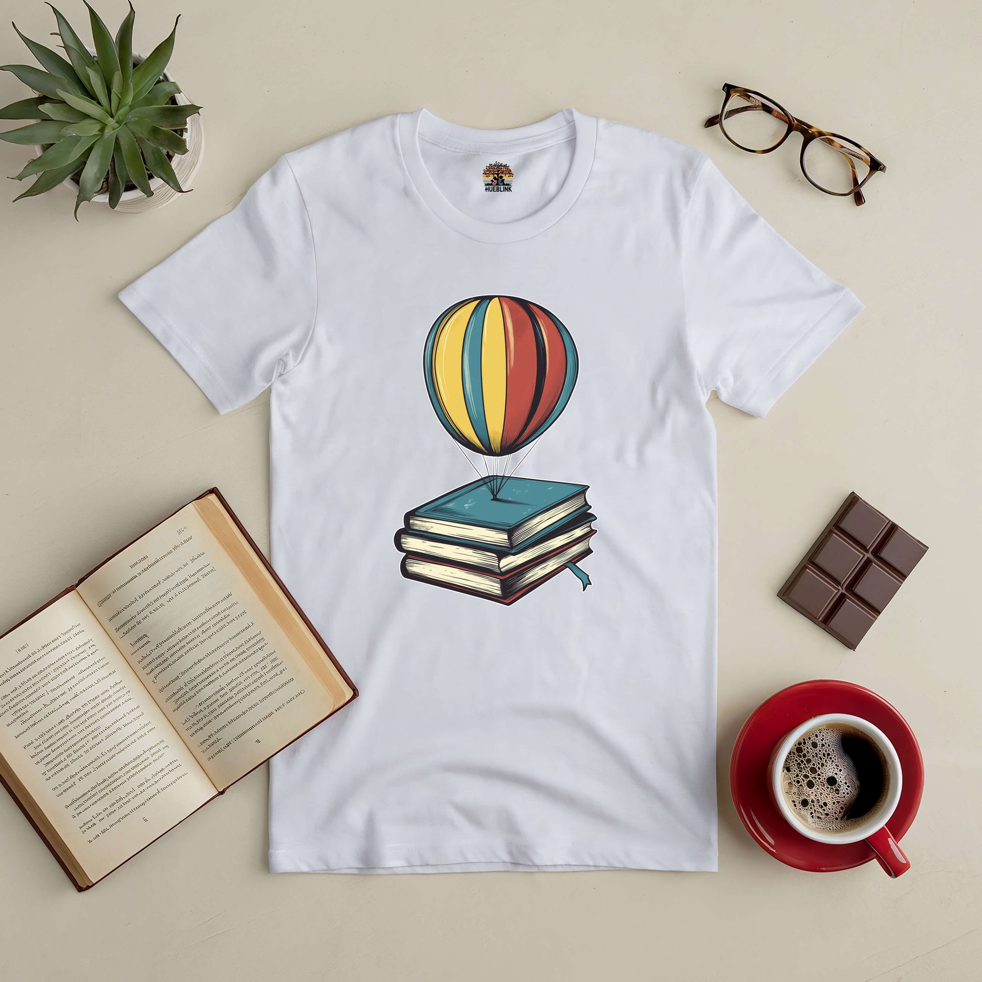 "Passport to Dreamland tee with hot air balloon and books design, surrounded by coffee, chocolate, glasses, and open book"