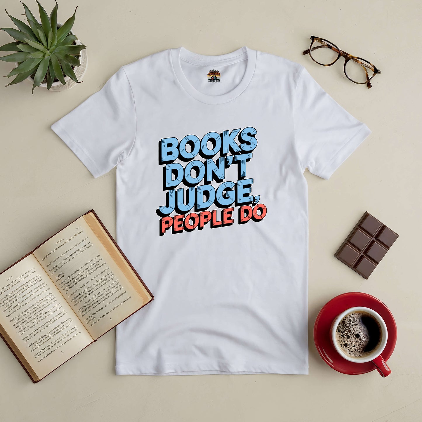 White tee with "Books Don't Judge, People Do" text, surrounded by an open book, coffee, chocolate, and glasses. Ideal for book lovers.