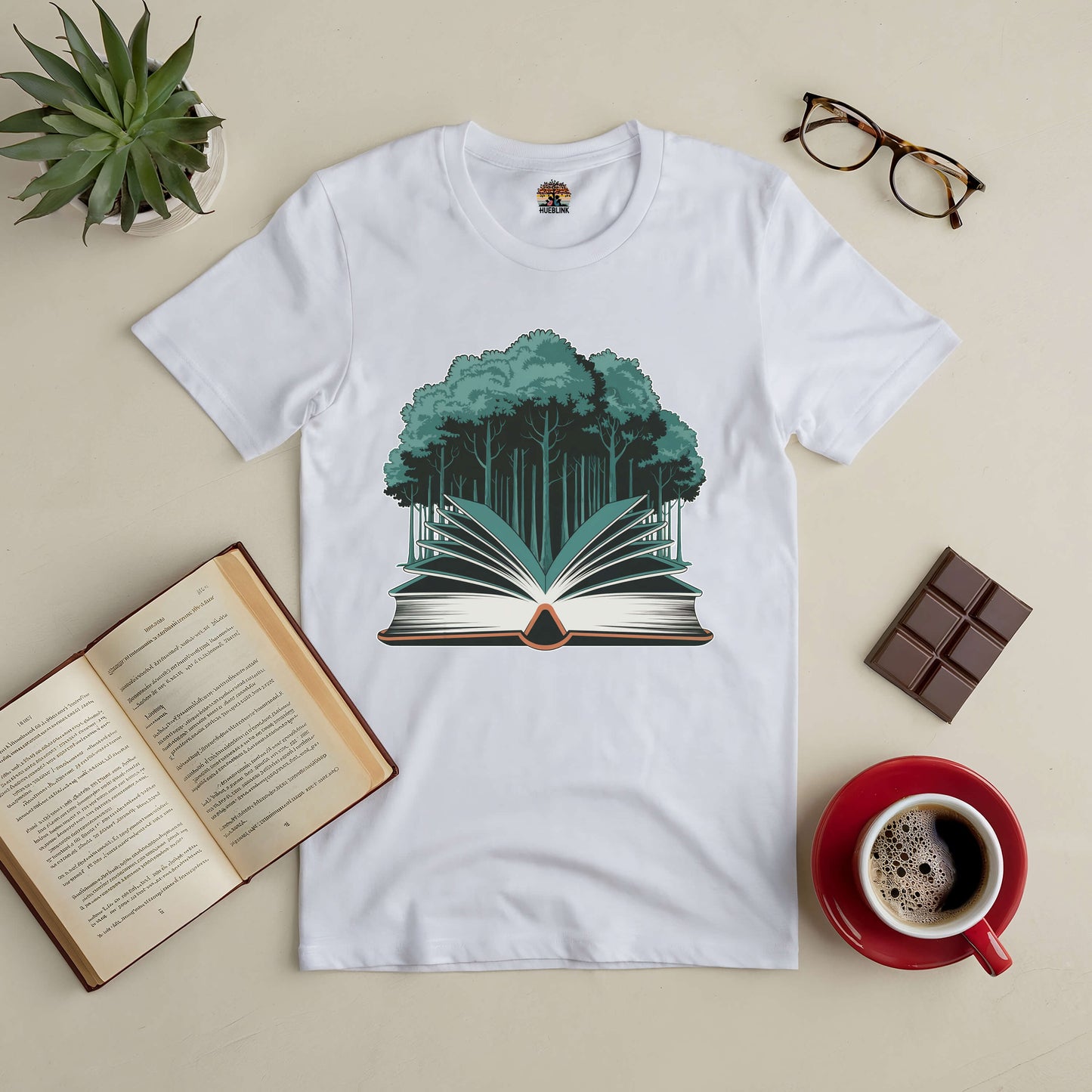 White t-shirt with forest and book graphic, surrounded by coffee, glasses, chocolate, plant, and an open book on a table.