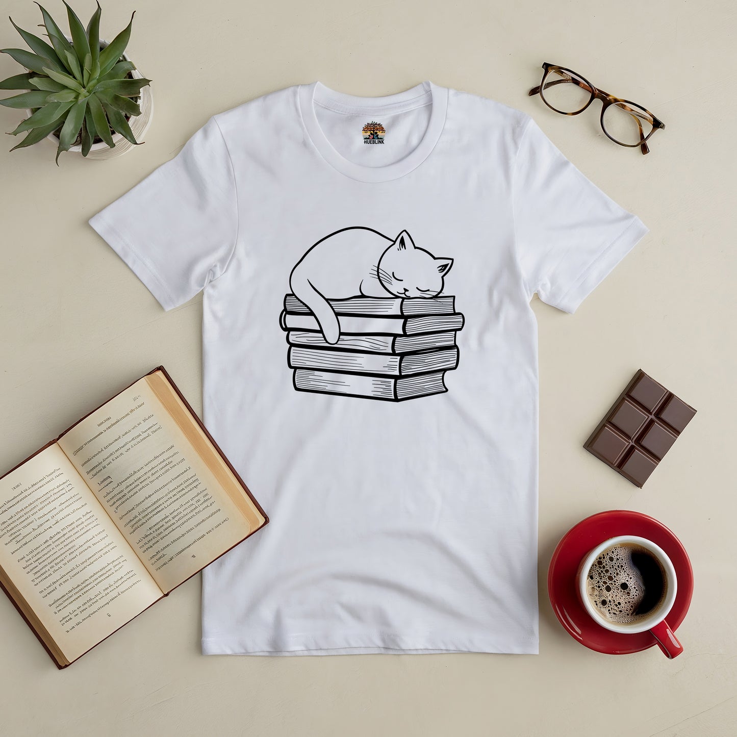 White tee with a cat sleeping on stacked books, surrounded by coffee, chocolate, and an open book for cozy book lovers.