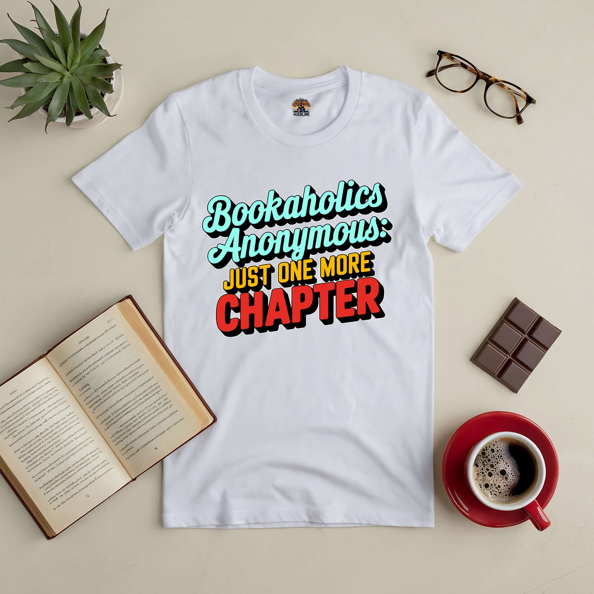 "White Bookaholics Anonymous Tee with 'Just One More Chapter' text, surrounded by a book, glasses, coffee, and chocolate bar."