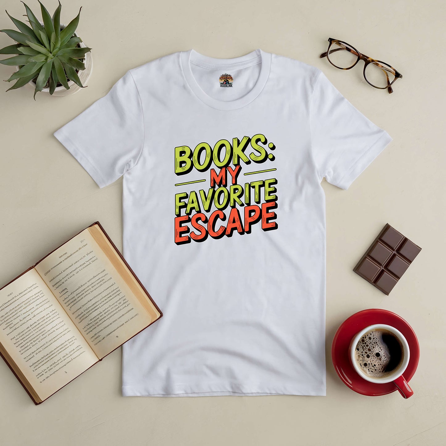 White "Books: My Favorite Escape" tee with open book, coffee, chocolate, glasses, and plant on a beige surface. Perfect for book lovers.