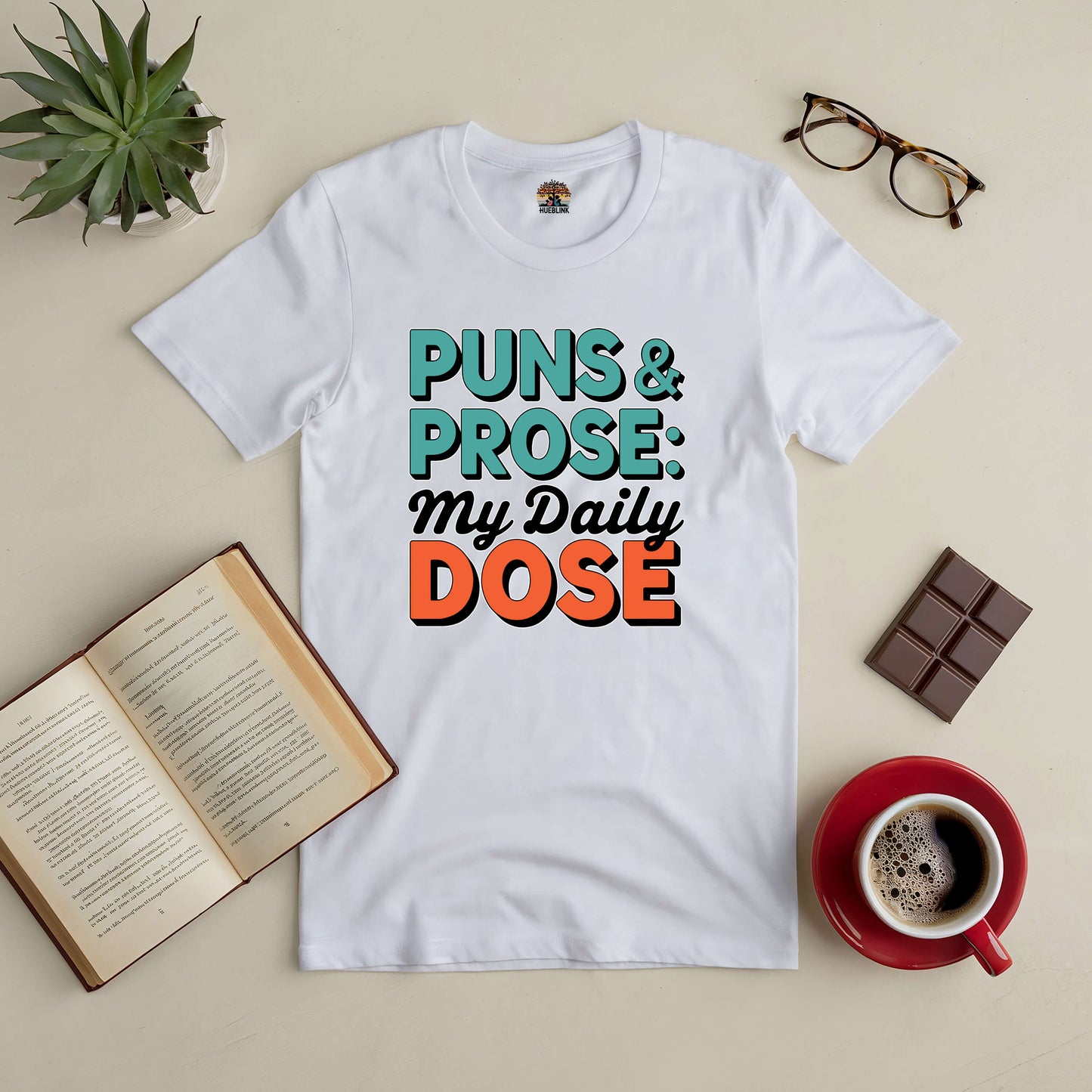 White tee with "Puns & Prose: My Daily Dose" text, surrounded by an open book, coffee, chocolate, glasses, and a plant.