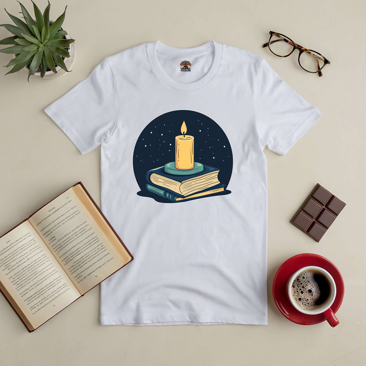 "Illuminate Your Mind Tee with candle and books design, surrounded by an open book, coffee, plant, glasses, and chocolate"