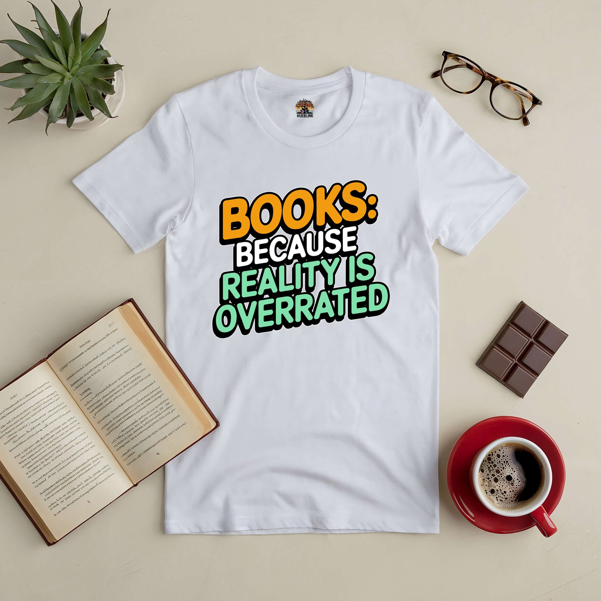 White t-shirt with "Books: Because Reality Is Overrated" text, displayed with an open book, glasses, coffee, chocolate, and a plant.