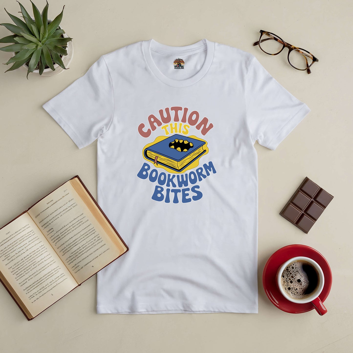 "Caution This Bookworm Bites tee with book graphic, surrounded by an open book, coffee cup, chocolate, and glasses on a table."