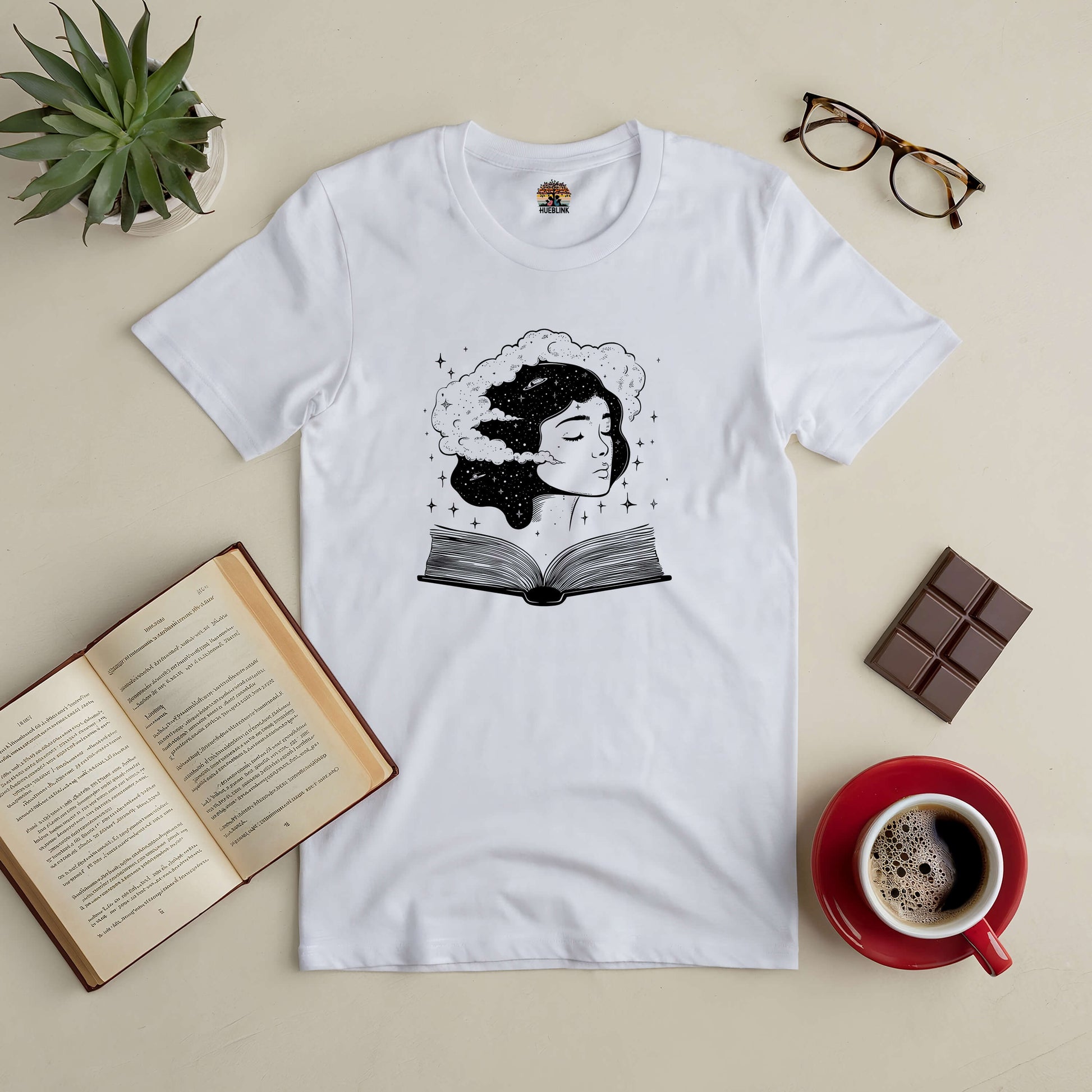 Universal Consciousness Tee with a cosmic design, surrounded by a book, coffee, plant, glasses, and chocolate on a table.