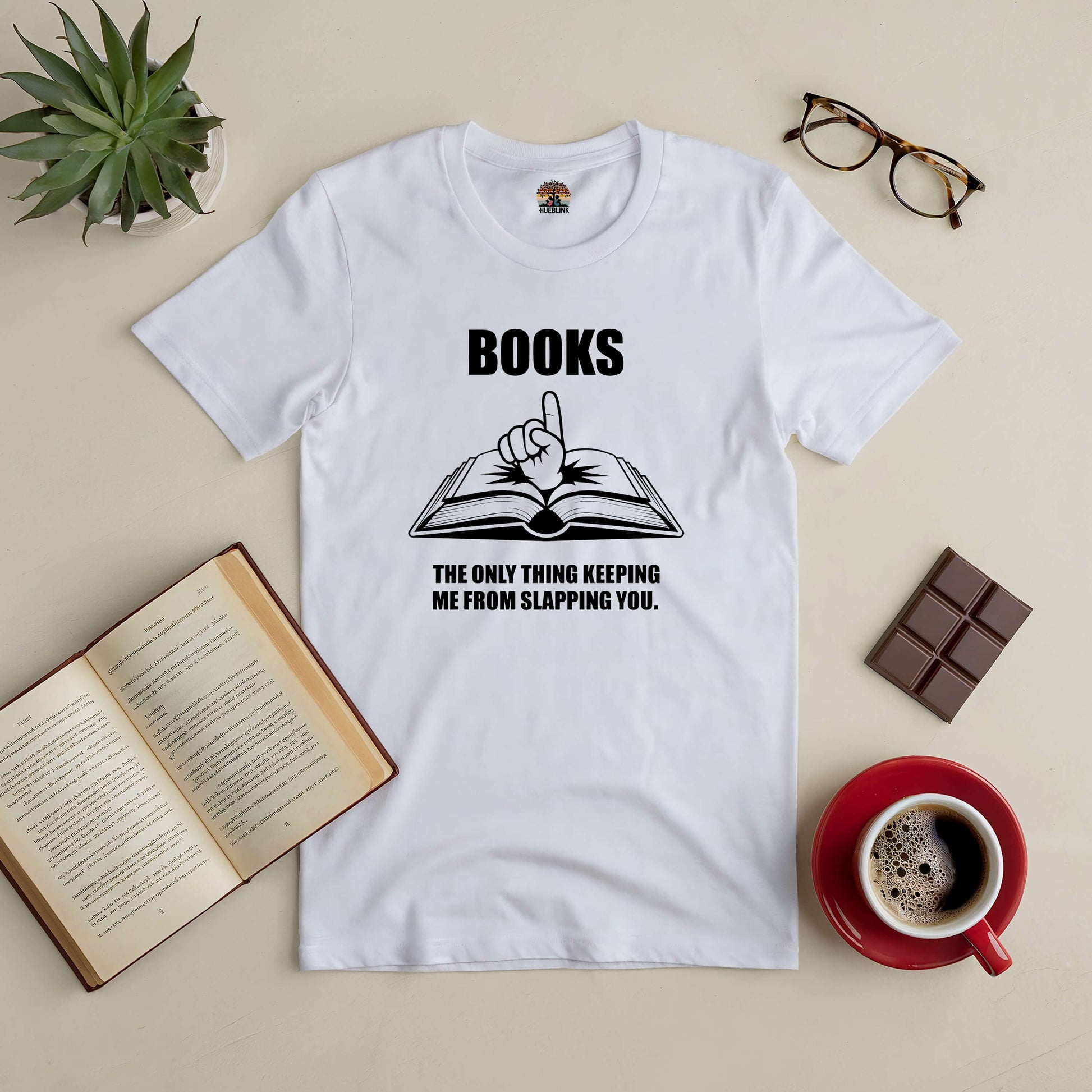 White tee with "Books: The Only Thing Keeping Me from Slapping You" text, styled with open book graphic, coffee, glasses, and plant.