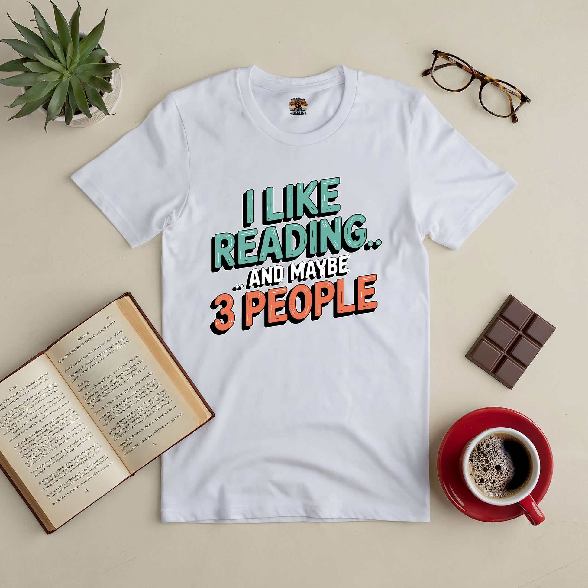 White tee with "I Like Reading... and Maybe 3 People" text, surrounded by an open book, glasses, chocolate, and coffee.