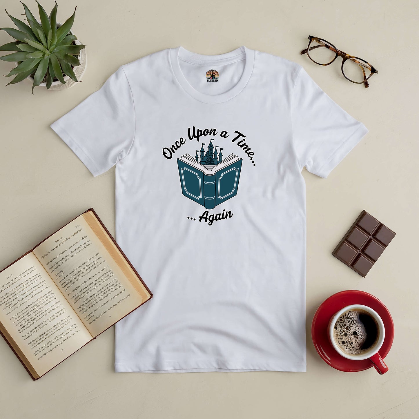 White "Once Upon a Time... Again" tee on a table with books, chocolate, glasses, and a coffee cup, perfect for dreamers and story lovers.