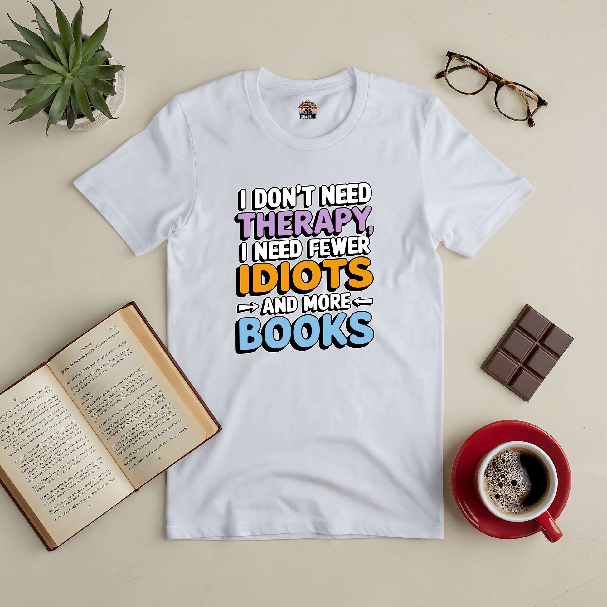 White tee with "I Don't Need Therapy, I Need Fewer Idiots and More Books" text, surrounded by coffee, glasses, and open book.