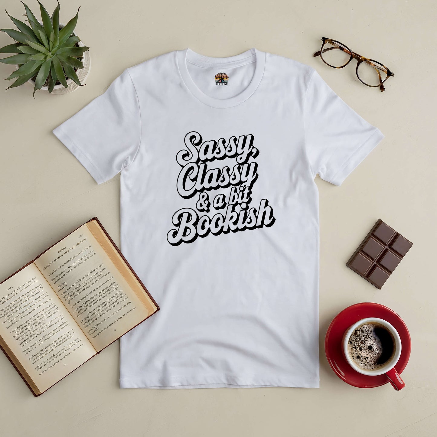 White Sassy, Classy & a Bit Bookish tee with a book, coffee, glasses, and chocolate bar on a table. Perfect for stylish book lovers.