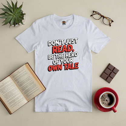 White tee with "Don't Just Read, Be the Hero of Your Own Tale" text, surrounded by glasses, coffee, chocolate, and an open book.