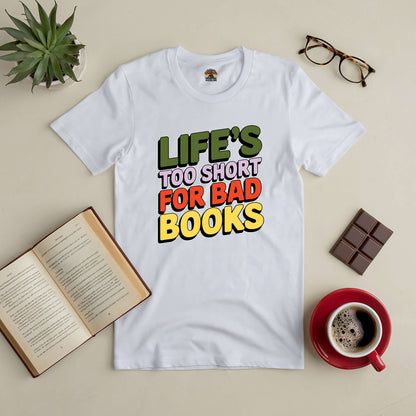 White tee with "Life's Too Short for Bad Books" slogan, surrounded by an open book, eyeglasses, chocolate, and coffee. Perfect for book lovers.