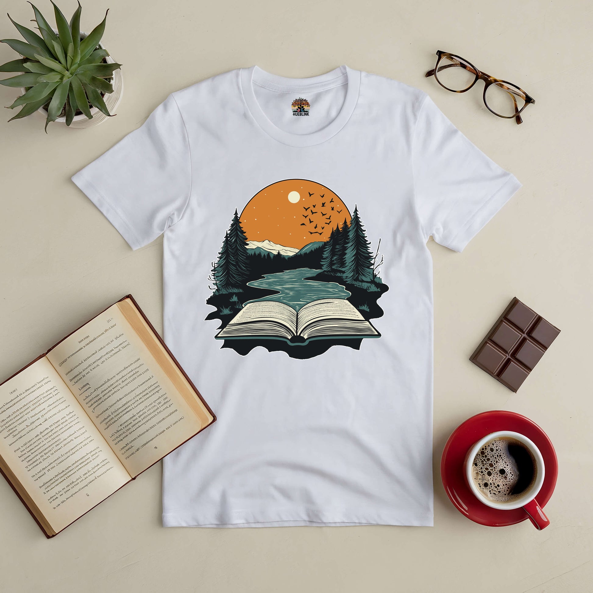 White t-shirt displaying a nature and book graphic, surrounded by an open book, coffee, plant, chocolate, and glasses.