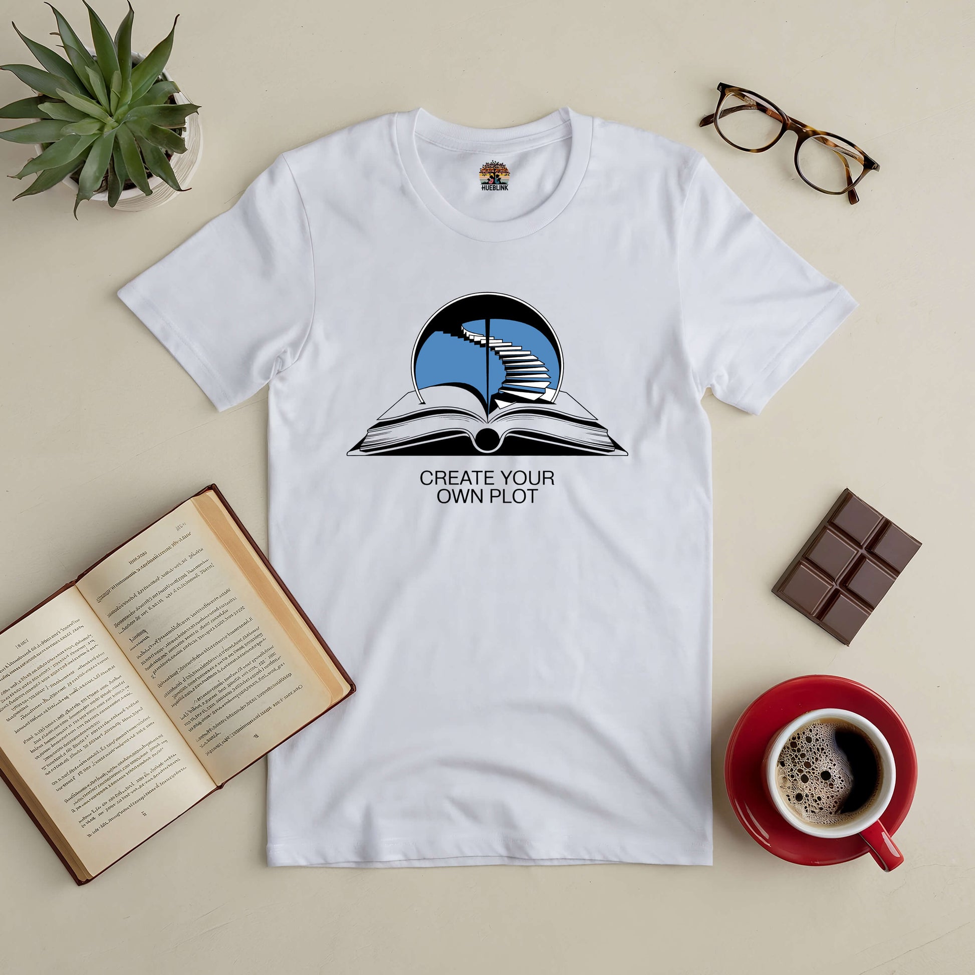 White tee with "Create Your Own Plot" design, featuring an open book and staircase, surrounded by coffee, glasses, plant, and chocolate.