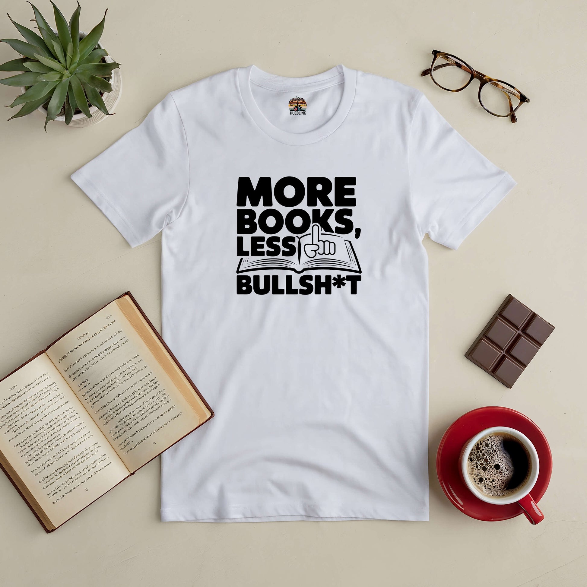 "White tee with 'More Books, Less Bullsh*t' slogan, surrounded by open book, coffee cup, chocolate, and glasses on a table"