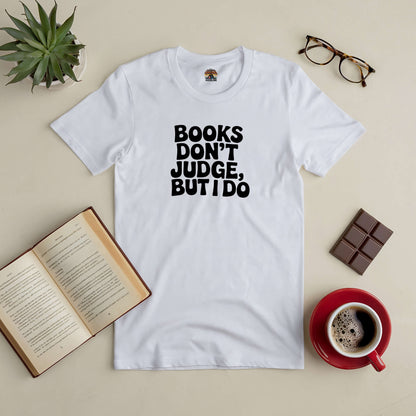 White tee with "Books Don't Judge, But I Do" text, perfect for book lovers, surrounded by an open book, glasses, coffee, and chocolate.