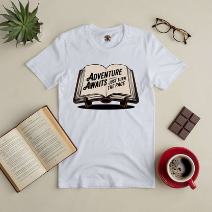 Adventure Awaits tee on a table with open book, coffee, plant, glasses, and chocolate; embrace new chapters stylishly.