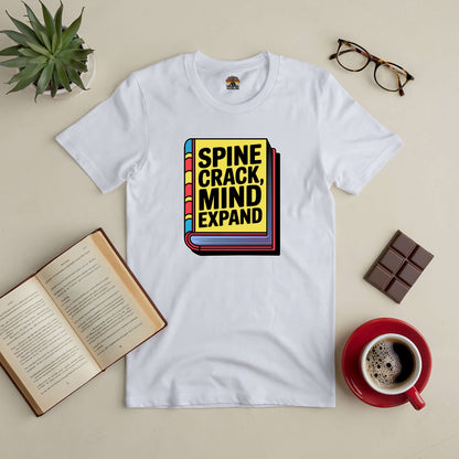 White "Spine Crack, Mind Expand" tee with book graphic, surrounded by open book, coffee, chocolate, glasses, and plant on table.