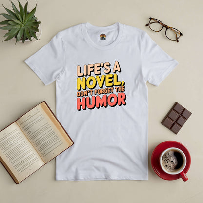 White tee with "Life's a Novel, Don't Forget the Humor" text, surrounded by glasses, coffee, a book, chocolate, and a plant.