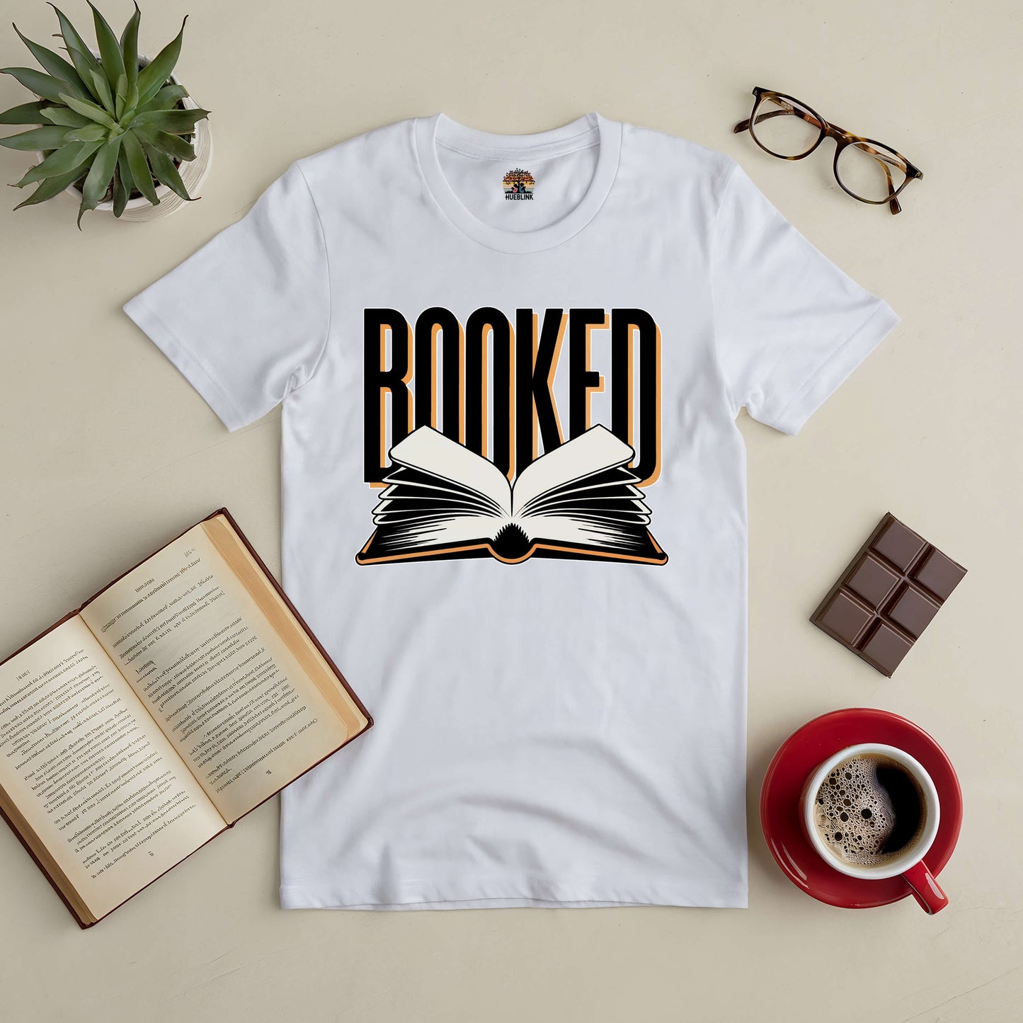 White "Booked" tee with open book design surrounded by an open book, coffee, glasses, and a succulent on a beige surface.