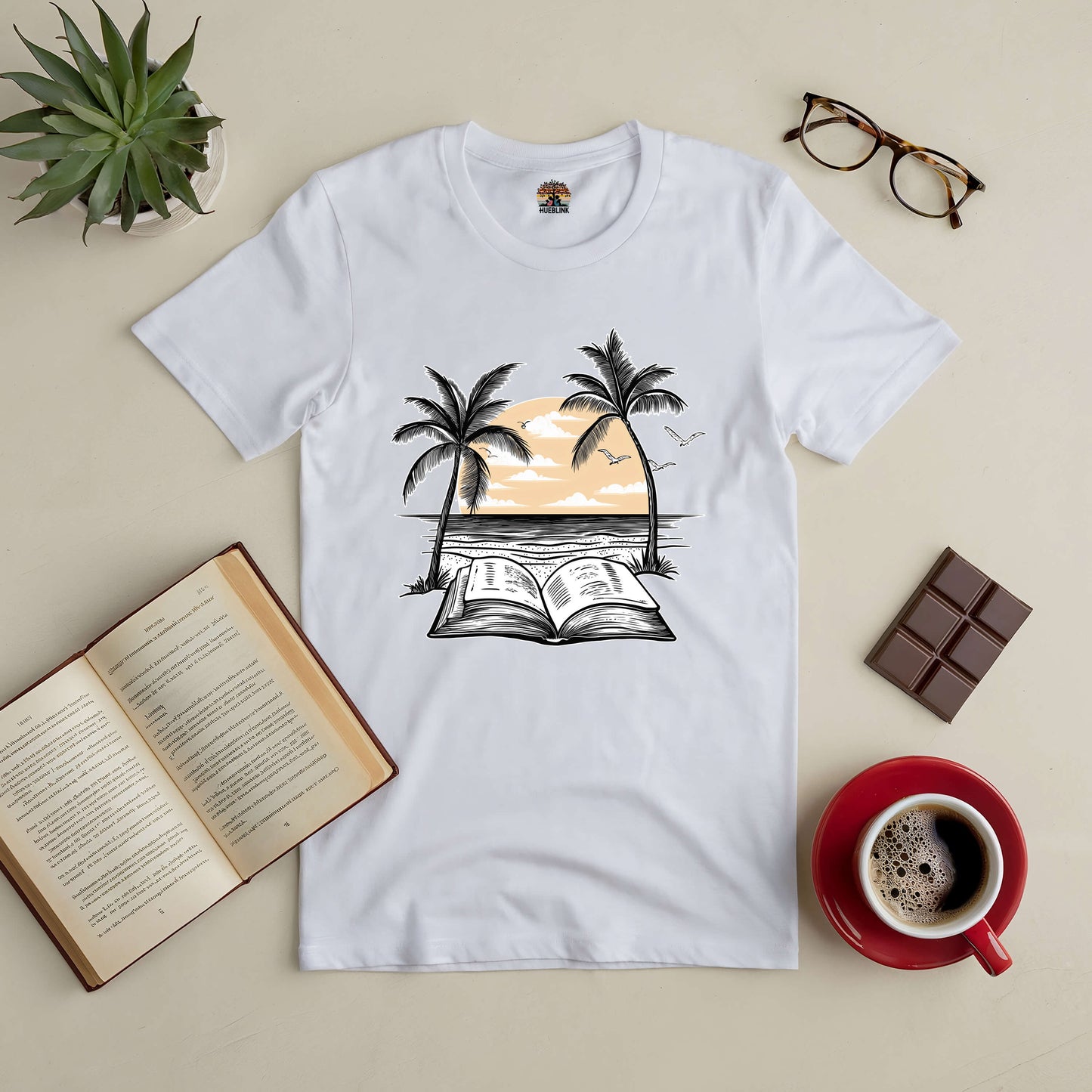 "Beachside Bibliophile Tee with palm trees, open book graphic, surrounded by coffee, chocolate, and open book on a beige table."