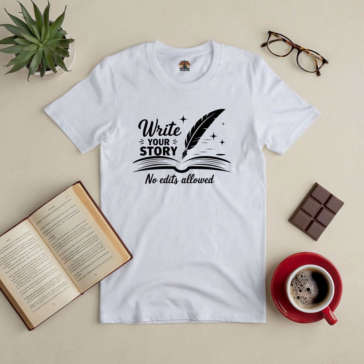 White tee with "Write Your Story, No Edits Allowed" design, surrounded by coffee, chocolate, book, glasses, and plant.