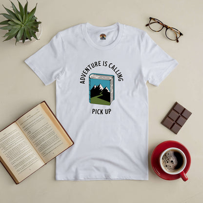 White t-shirt with "Adventure Is Calling, Pick Up" design, surrounded by an open book, glasses, plant, chocolate, and coffee cup.